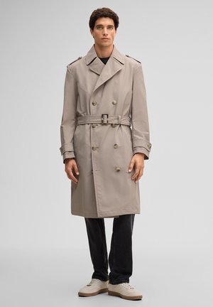Uomo che indossa un trench beige doppiopetto con cintura, pantaloni neri e sneakers bianche, in piedi davanti a uno sfondo grigio chiaro uniforme.