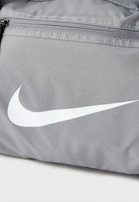 Šedá sportovní taška z texturované tkaniny s velkým bílým logem Nike ve tvaru křídel a kapsou na zip.