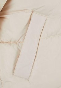 Polstret vest i lys beige stoff, med en glatt tekstur og en rektangulær lappdetalj på den ene siden. Synlige sømmer og kant.