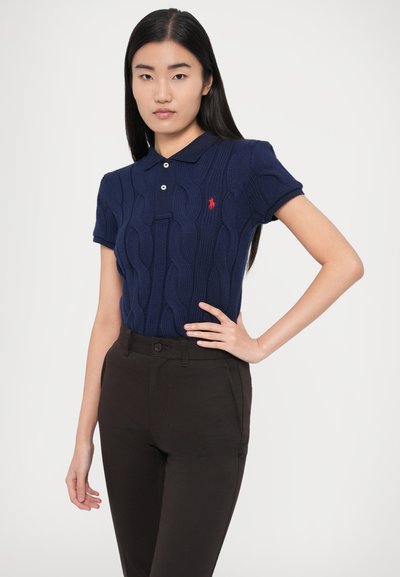 Μπλε ναυτικό πουλόβερ πικέ πόλο με γιακά και δύο κουμπιά, στολισμένο με ένα μικρό κόκκινο λογότυπο. Συνδυασμένο με σκούρο καφέ εφαρμοστά παντελόνια.