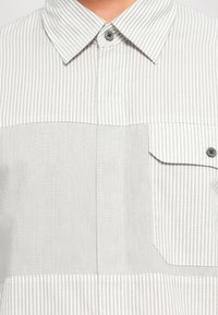 Gros plan d'une chemise blanche à rayures grises verticales avec un col pointu, un bouton supérieur fermé et une poche poitrine boutonnée côté droit.