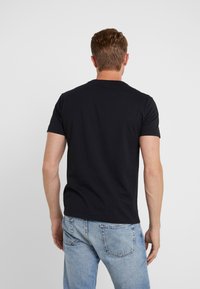 Muž stojící s otočením zády, oblečený v jednoduchém černém tričku s krátkým rukávem a světle modrých džínách proti jednoduchému pozadí.