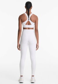 Bijeli sportski crop top s dizajnom križanja na leđima i izrezom, u kombinaciji s bijelim tajicama viskog struka koje imaju ravne šavove.