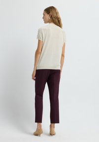 Gebreide top met korte mouwen in crème met een gestructureerde schouderaccenten, gecombineerd met bordeauxrode broek en beige schoenen, gezien van achteren.