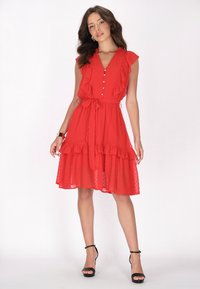 Rotes Kleid aus leichtem Stoff, mit einem Volantdesign, Knopfschluss und einem Gürtel in der Taille. Schwarze sandalen mit Riemchen werden getragen.