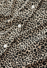 Camisa de estampado de leopardo en beige y negro, con cuatro botones blancos, textura acanalada y un patrón animal distintivo en todo su diseño.