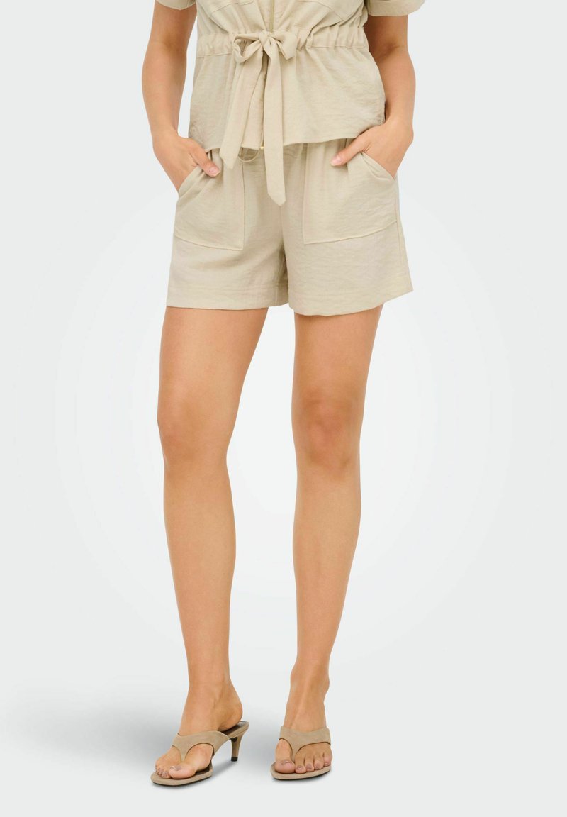 Haut beige noué à la taille et short assorti avec poches devant, porté avec des sandales à talons beige sur une personne debout.