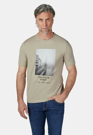 Mittelalter Mann mit grauen Haaren, der ein beiges T-Shirt mit schwarz-weißem Paris-Aufdruck und blaue Jeans trägt und mit einer Hand in der Tasche steht.