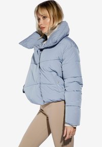 Giacca imbottita blu chiaro con colletto alto, chiusura con zip e tasche laterali. Il design trapuntato aggiunge consistenza e dimensione.