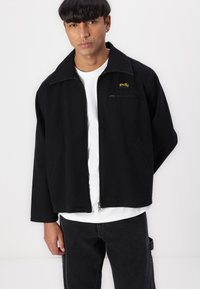 Svart zip-up jakke laget av bomull, med spiss krage, sidelommer og en liten gul logo på venstre bryst. Brukes over en hvit skjorte.