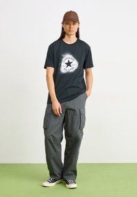 Juoda marškinėliai su balta Converse logotipu, pilki kargo kelnės, rudos spalvos kepurė ir juodi sportiniai bateliai su baltais padu. Paprasta, laisva forma ir dizainas.