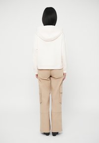 Oseba z ravnimi črnimi lasmi nosi belo kapuco in bež cargo hlače, stoji obrnjena stran od preproste bele ozadja.