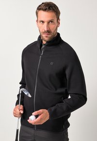 Giacca nera con zip e colletto alto, realizzata in tessuto liscio. Modello che tiene in mano una mazza da golf e una pallina, con dettagli del logo in modo discreto sul petto.