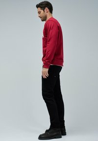 Sweat-shirt rouge avec texte blanc, pantalon noir et bottes noires, se tenant contre un fond neutre. Vue de profil.