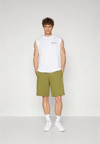 Sweatshirt branco sem mangas com logótipo, combinado com calções verde-oliva. Apresenta um ajuste descontraído e ténis brancos com meias texturizadas caneladas.