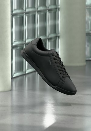 Czarny skórzany sneaker ze sznurowadłami i logo "BOSS", unoszący się nad refleksyjną betonową podłogą w nowoczesnym wnętrzu z ścianą z bloków szklanych.