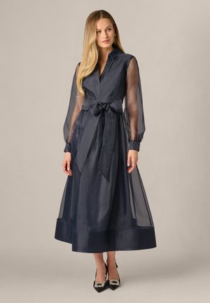 Kobieta ubrana w granatową przezroczystą sukienkę midi z długim rękawem, kołnierzykiem, wiązaniem w talii i czarne, szpiczaste szpilki z ozdobnymi klamrami.