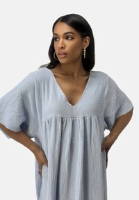 Robe bleu clair, ample, en tissu texturé avec manches larges, col en V et taille froncée. Pas de motifs ni de détails visibles.