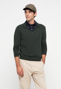 Uomo che indossa un maglione verde scuro con zip a quarti sopra una polo blu navy, pantaloni beige e un cappello piatto a scacchi, in piedi con una mano in tasca.