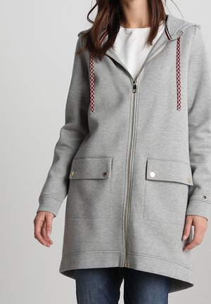 Frau trägt einen langen grauen Zip-Hoodie mit zwei vorderen Klappentaschen und rot-weißen Kordeln über einem weißen Shirt und blauer Jeans.