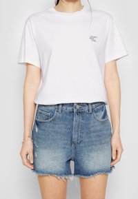 Osoba oblečená v bílem tričku s nápisem "CRAZY SEXY COOL" a v potrhaných modrých džínových šortkách s vysokým pasem a roztrhanými okraji.