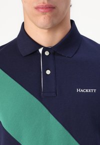 Marineblå polo med grønn diagonal stripe, to knapper ved kragen og "Hackett"-logo brodert i hvitt. Teksturert stoff.