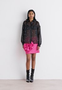 Schwarze durchsichtige Bluse mit weißen Verzierungen, kombiniert mit einem rosa Minirock mit einer großen Schleifenapplikation und schwarzen Ankle Boots.