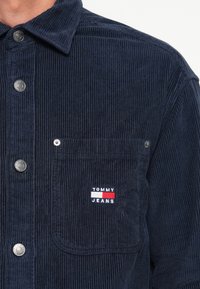 Jūros mėlynos spalvos švarkas iš kordūro su metaliniais mygtukais, pasižymintis krūtinės kišenėle ir išsiuvinėtu "TOMMY JEANS" logotipu raudonos, baltos ir mėlynos spalvų.