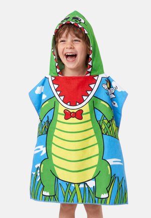 Kind lächelt und trägt ein farbenfrohes Kapuzenhandtuch, das als grüner Cartoon-Krokodil mit roter Fliege gestaltet ist, und eine Outdoor-Kulisse.