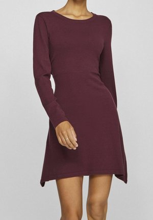 Femme portant une robe ajustée bordeaux foncé à manches longues, longueur genou, avec un col rond, sur un fond gris uni.