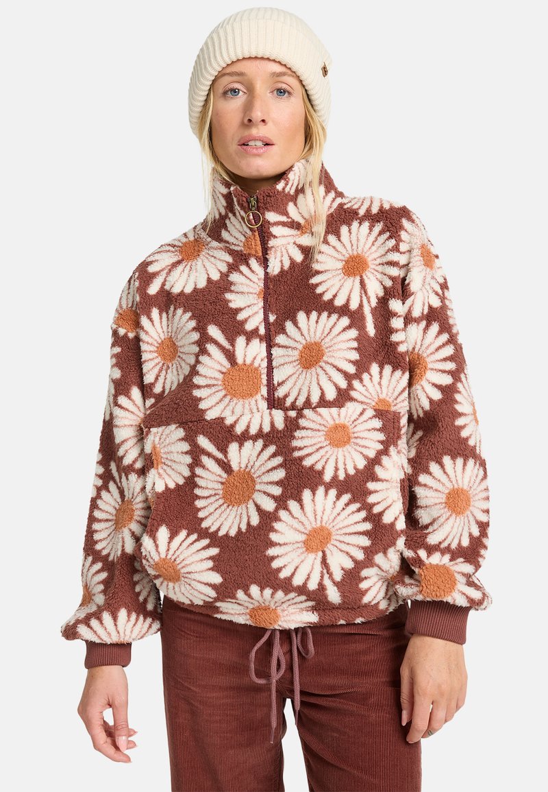 Brun fleece-pull med vitt och orange prästkrage-mönster, halvdragkedja fram, elastiska ärmslut och dragsnöre i nederkanten. Bärs med en krämfärgad mössa.