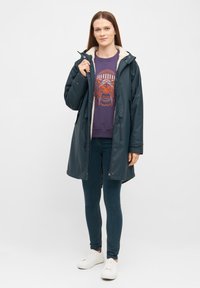 Parka impermeabile verde scuro con cappuccio foderato in pelliccia, chiusura con zip e lacci. Indossato sopra una felpa viola con un design grafico arancione.