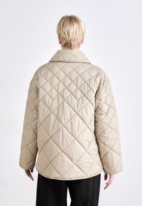 Μπεζ καπιτονέ ζακέτα με διαμαντένιο σχέδιο, φαρδύ γιακά και άνετη εφαρμογή. Υφασμάτινη υφή και μακριά μανίκια. Χωρίς ορατά αξεσουάρ ή λεπτομέρειες.