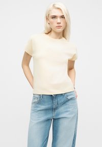 Νέα γυναίκα με μακριά ξανθά μαλλιά, φορώντας απλό κρεμ κοντομάνικο πουκάμισο και ανοιχτόχρωμα μπλε τζιν ψηλόμεσα, με τα χέρια στις τσέπες, ουδέτερη έκφραση.