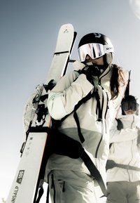 Ski gear with a white and light gray insulated jacket, black gloves, and mirrored goggles; skis carried on the back; bright sky backdrop.