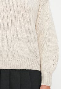 Kremowy, dziany sweter o luźnym kroju, z prążkowanym dołem i szerokimi rękawami. Unikalna struktura dzięki wyraźnym ściegom. Połączony z ciemną spódnicą.