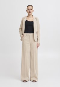 Beige přizpůsobený blazer a široké kalhoty, texturovaná tkanina, hladký černý top pod ním, s minimalistickým designem a střihem.
