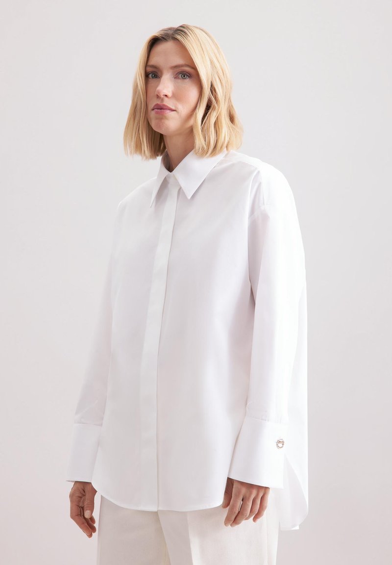 Femme aux cheveux blonds mi-longs portant une chemise blanche surdimensionnée à boutons avec de longues poignets et un pantalon blanc sur un fond uni.
