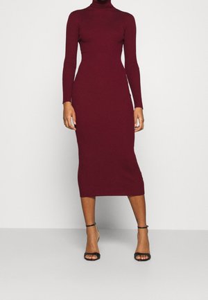 Robe en maille côtelée bordeaux avec col haut et manches longues, silhouette ajustée s'évasant jusqu'à mi-mollet. Talons noirs à lanières.