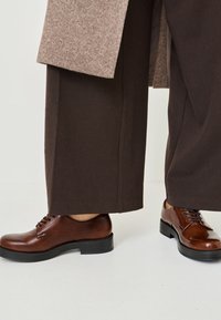 Zapatos oxford de cuero marrón con textura suave, punteras redondas y suelas de goma negra, combinados con pantalones marrones y un abrigo estampado.