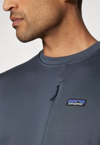 Navy blue long-sleeve top with a round neckline, featuring a small Patagonia logo patch and a decorative drawstring on the shoulder.