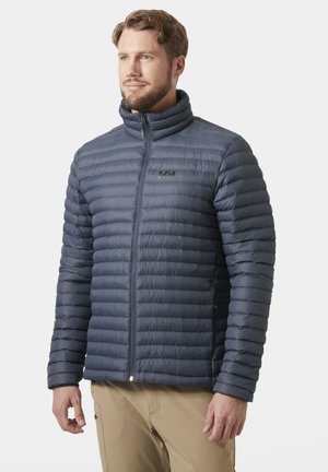 Hombre con chaqueta acolchada azul marino con cremallera y pantalones beige, de pie y mirando ligeramente de lado sobre un fondo liso.