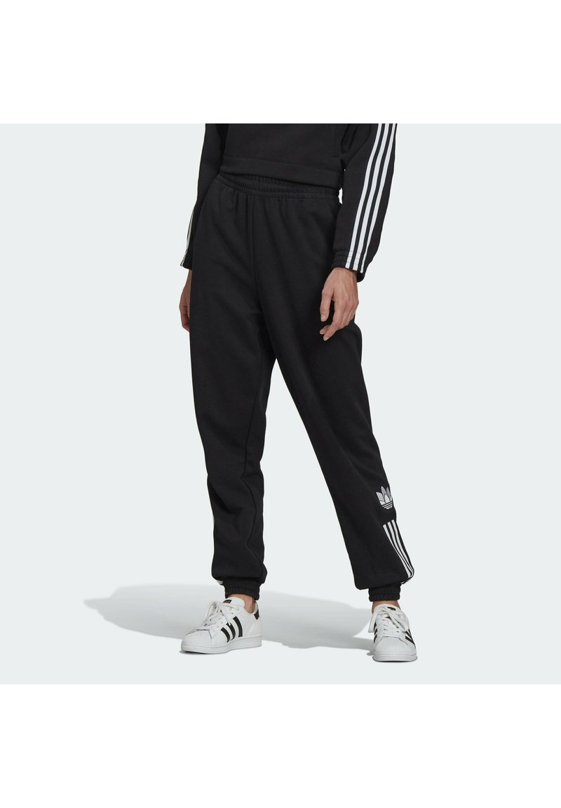 Person som bär svarta Adidas-joggingbyxor med vita ränder och logotype, svart sweatshirt med vita ränder på ärmarna, och vita Adidas-sneakers.