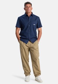 Marineblaues Kurzarmhemd mit Brusttasche und dezentem Logo, kombiniert mit lockeren beigen Hosen und weißen Sneakers. Glatte Stofftextur.