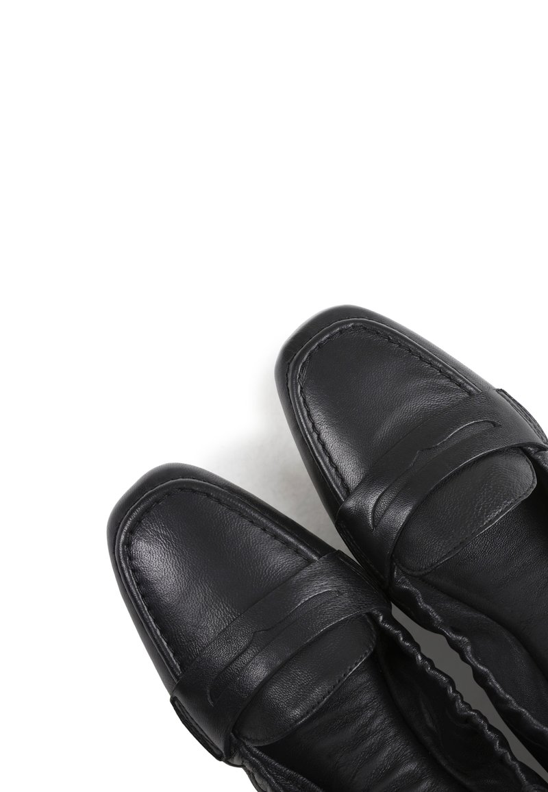 Zwarte leren loafers met een vierkante neus en een banddetail over de bovenkant, tegen een witte achtergrond.