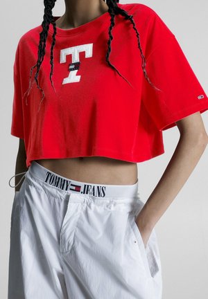 Osoba nosząca czerwony krótki T-shirt z dużą białą literą T i białe spodnie z widocznym pasem Tommy Jeans.