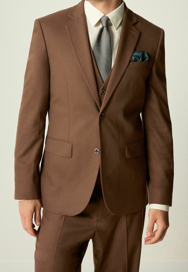 Homme portant un costume trois pièces marron avec une chemise blanche, une cravate grise à motifs et un mouchoir de poche vert foncé, debout les bras relâchés.
