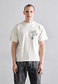 Kremfarget bomulls-T-skjorte med en grå grafikk som sier "MOTEL GOD" i et korsdesign på forsiden. Korte ermer, avslappet passform, minimal tekstur.