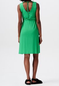 Vestido verde de longitud hasta la rodilla, hecho de una tela suave, con un escote en la espalda y un detalle de lazo. Llevado con sandalias negras, mostrando una textura suave y un ajuste relajado.