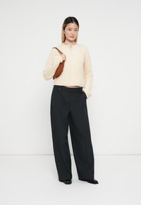 Cardigan crem cu model în dungi, cu închidere cu nasturi, asortat cu pantaloni negri cu dungi largi și o geantă maro. Pantofi negri completează ținuta.